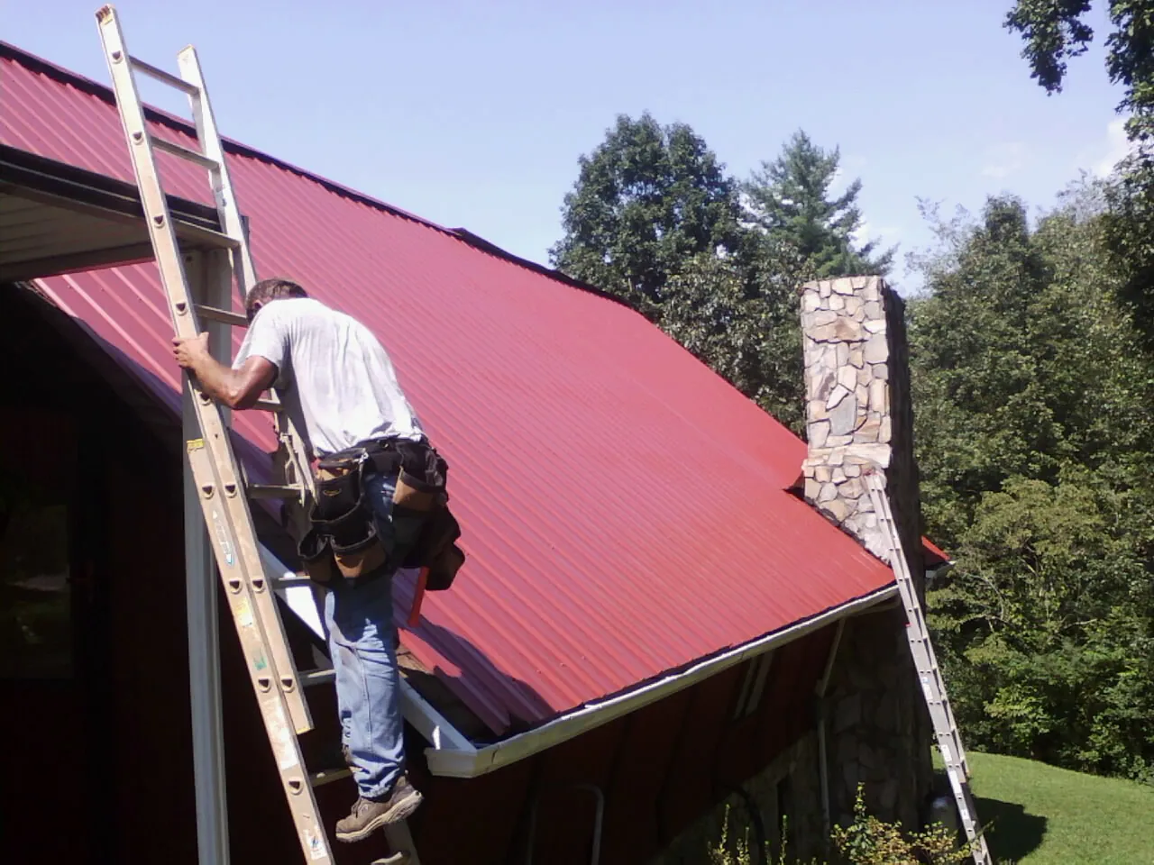 Roofer on a ladder beside a red metal roof and stone chimney — Soffit & Fascia Repair by Roofing Brothers in Calumet